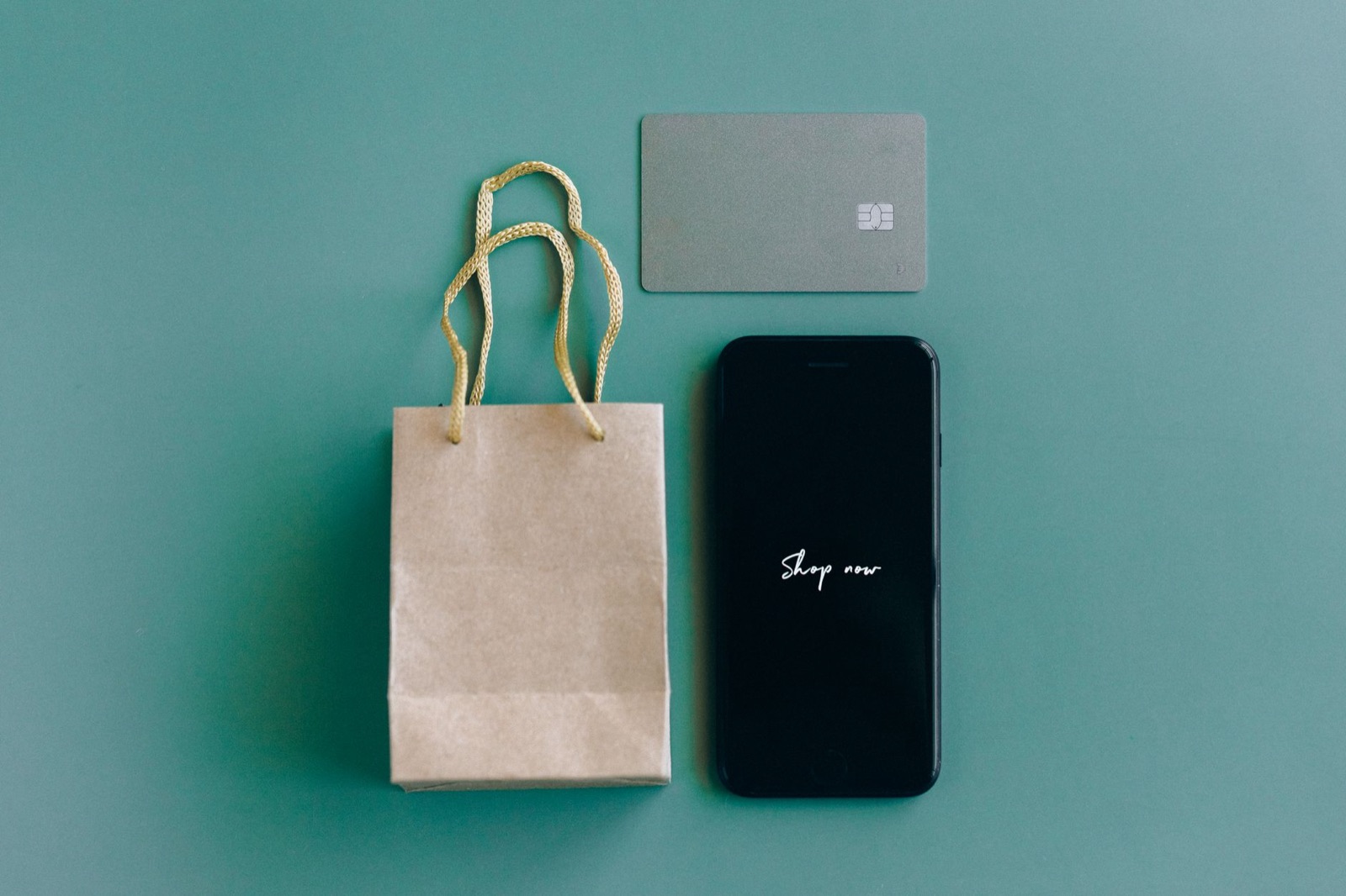 Flat lay on a teal background: a small kraft paper gift bag with cord handles, a plain grey chip card, and a phone whose screen reads Shop now.