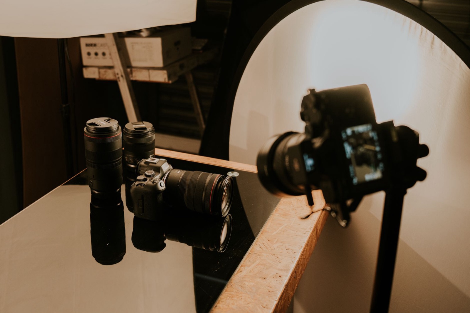 A camera on a stand shooting a row of lenses arranged on a lit tabletop set, with a curved white backdrop and a softbox overhead