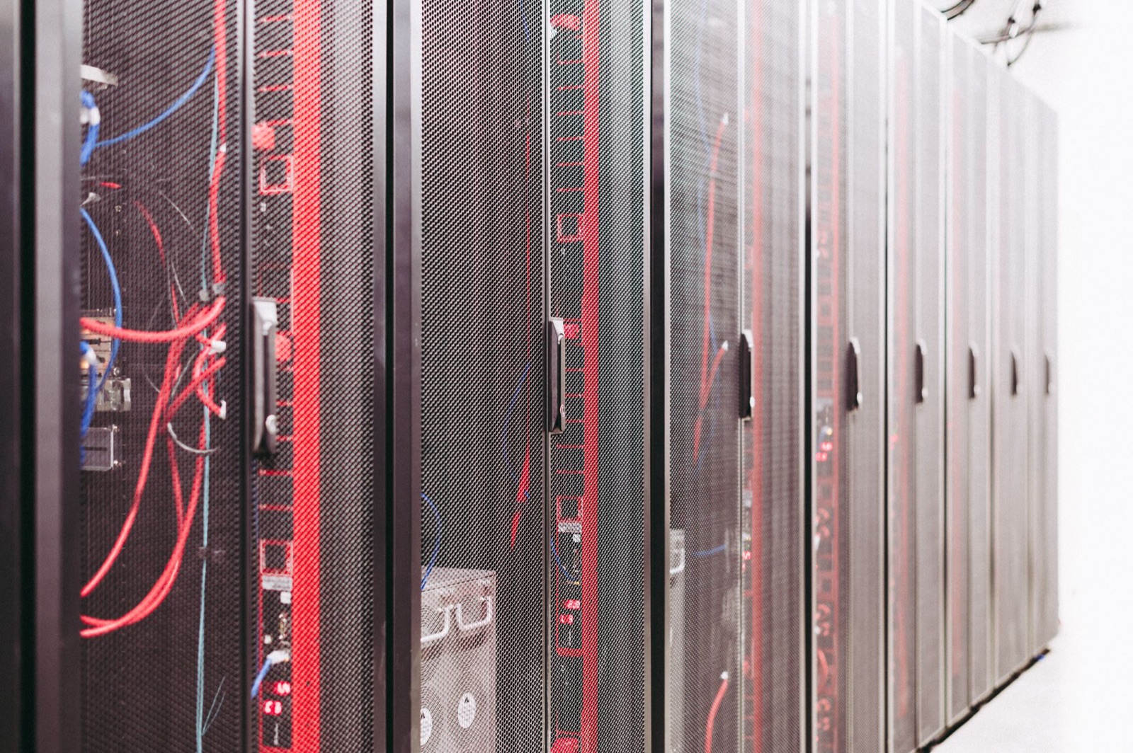 A row of black server racks in a data centre, mesh front doors closed, with red and blue patch cables visible inside the nearest cabinet.